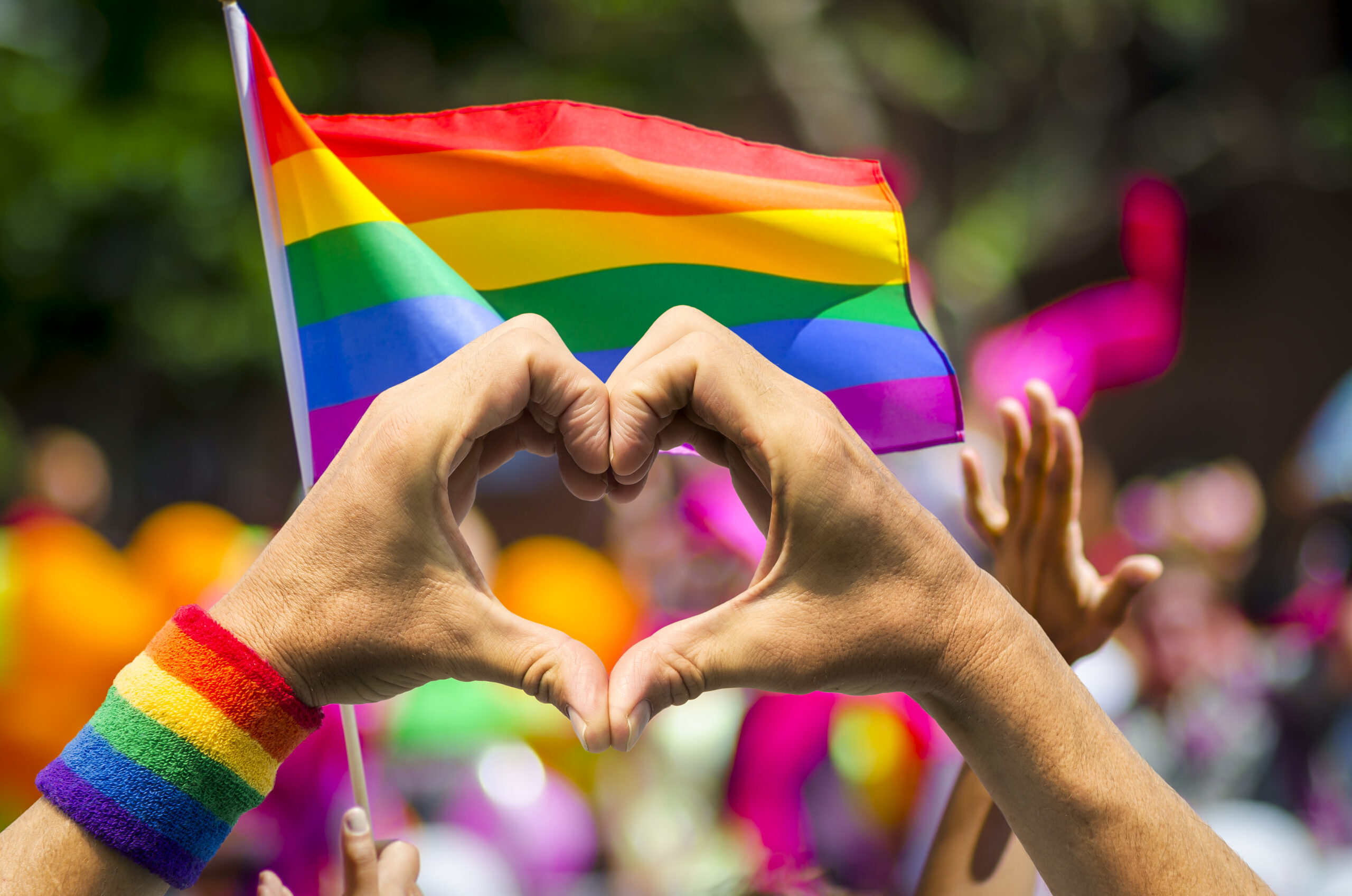 Image of a person's hands forming a heart in front of a rainbow flag