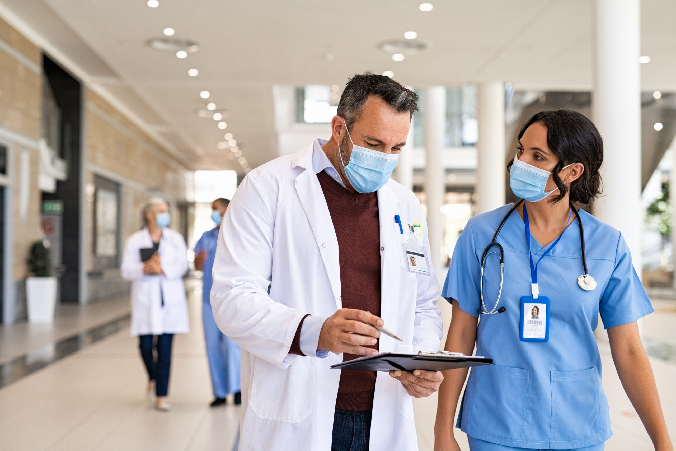Two healthcare professionals walk alongside one another. One is wearing a white coat, and the other has on blue scrubs. 