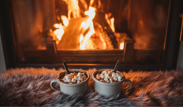 An image of two cups of hot cocoa in front of a fireplace