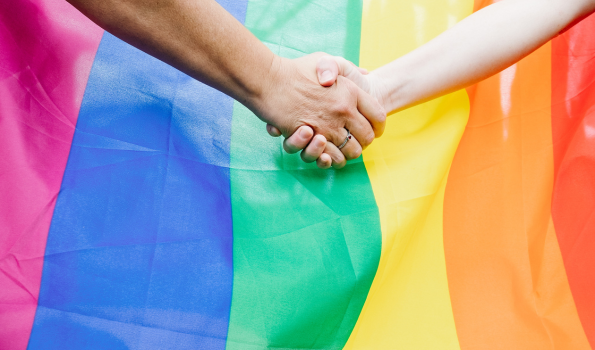 Two hands clasp together in front of a Pride flag. 