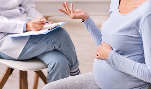 A pregnant person touches their stomach while speaking to someone holding a clipboard. 