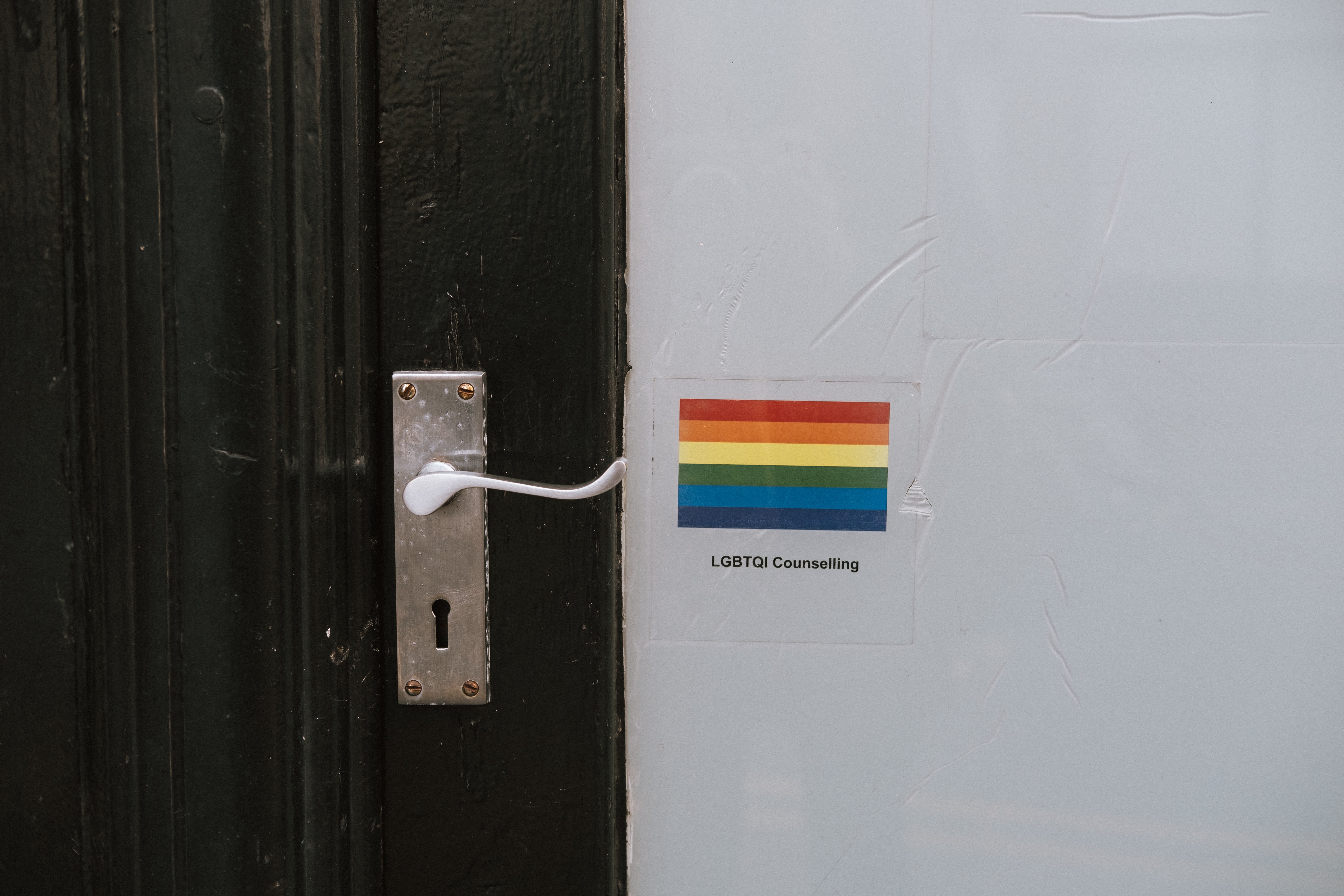 A pride rainbow flag sticker on a wall next to a wooden door with a silver door handle. Below the stick are the words "LGBTQI Counselling"