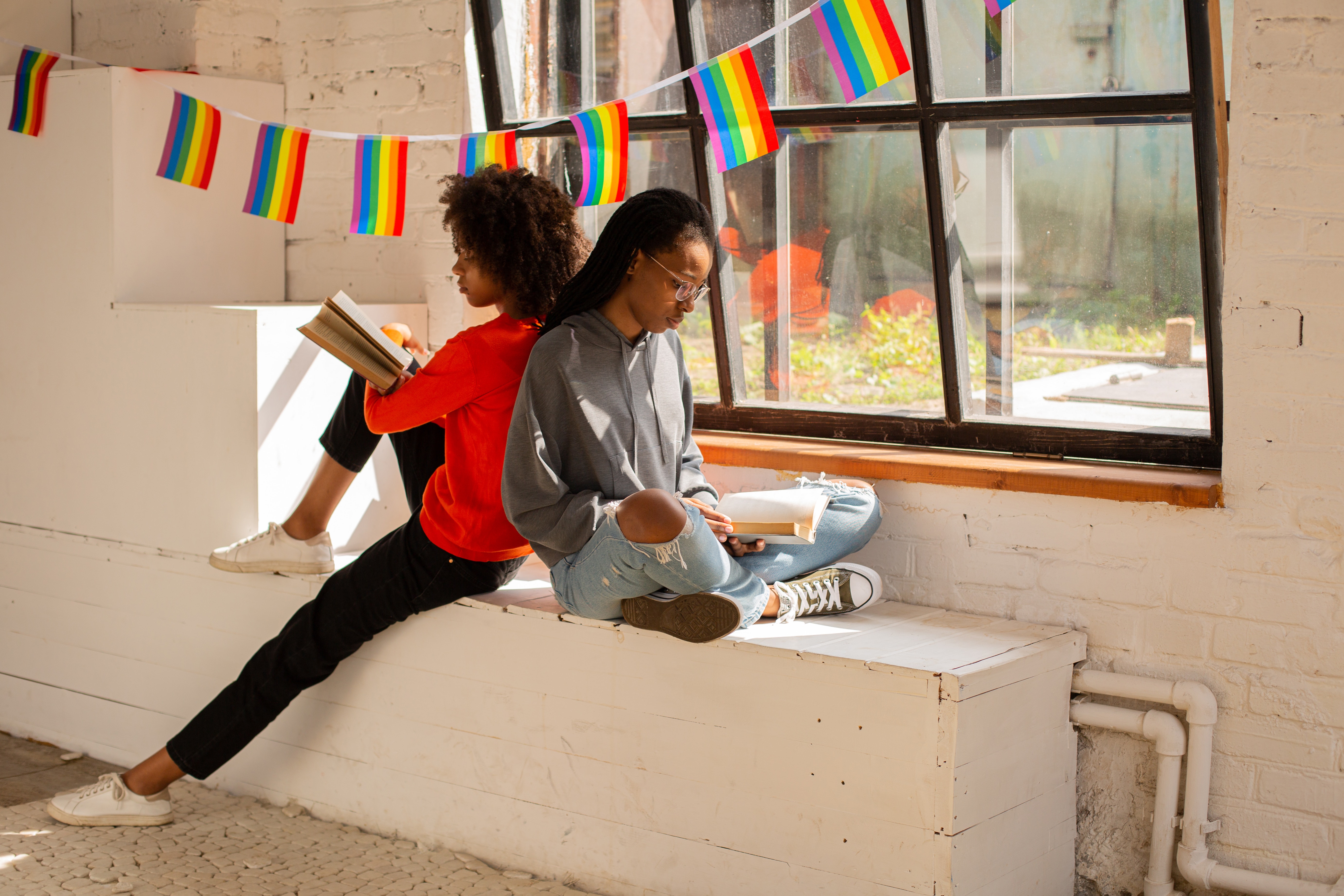Two youth in a classroom setting, reading books while sitting back to back with each other. Above the youth is a banner with small pride flags hung in the window of the classroom. 